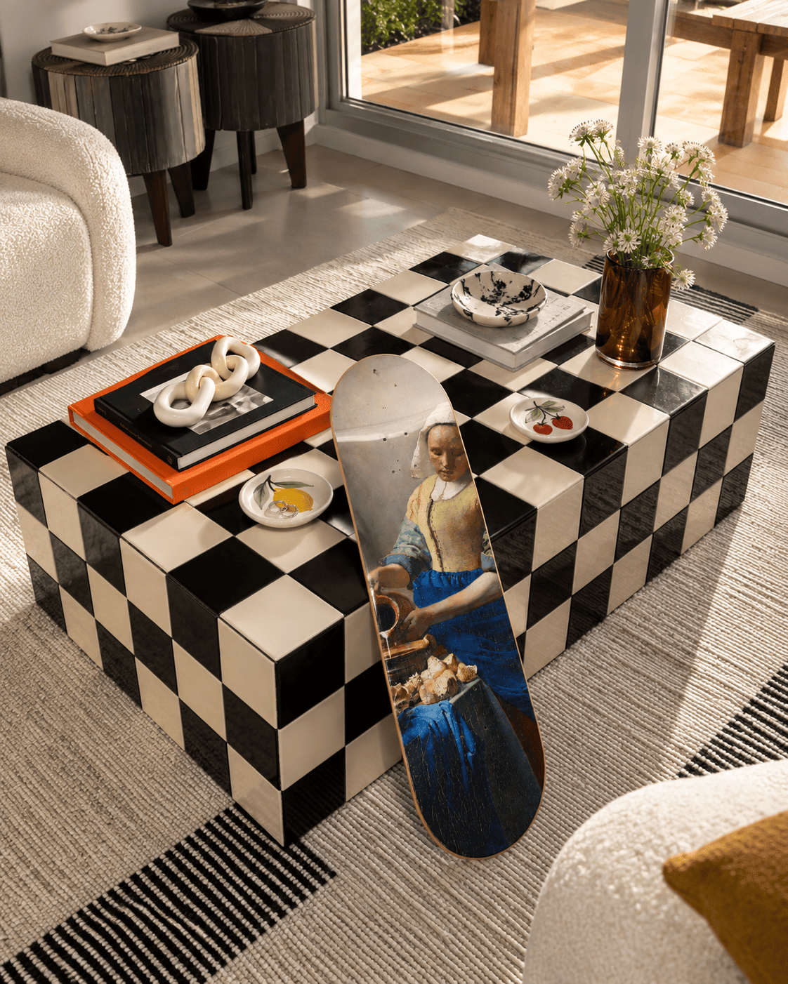 Vermeer's The Milkmaid on a skateboard deck leaning against a black-and-white checkerboard coffee table in a styled living-room scene with a bouclé chair, wildflowers and a large window